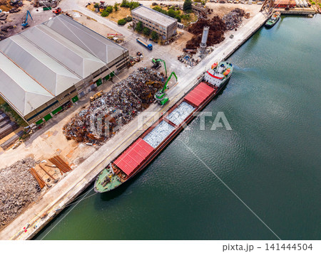 Ship docked at a warehouse with scrap metal and machinery during a sunny day in an industrial area by the water Ship docked at a warehouse with scrap metal and machinery during a sunny day in an industrial area by the water 141444504