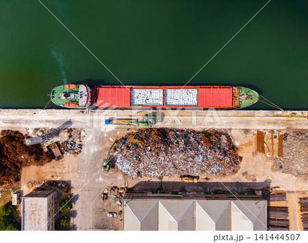 Cargo ship docks at port to unload materials near recycling area along the water during daytime Cargo ship docks at port to unload materials near recycling area along the water during daytime 141444507