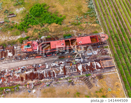 Rusty ship abandoned in a field near railway tracks and scattered debris in a rural area during a sunny day 141444509