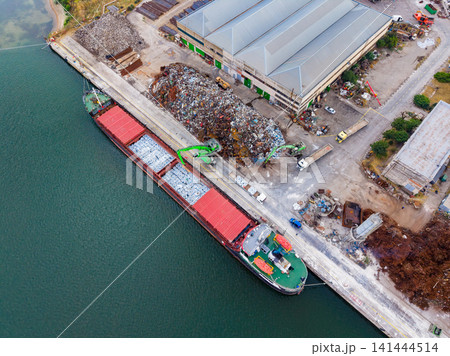 Cargo ship docks at port to unload materials near industrial site with large piles of scrap and storage buildings in the background during day 141444514