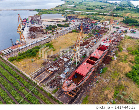 Ships sit at a dock with cranes nearby in a harbor area where boats are dismantled and scrap metal is processed during the day 141444515