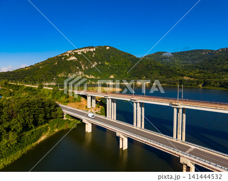Bridges cross over a river with a truck traveling on a highway surrounded by hills and clear blue skies in a sunny setting 141444532