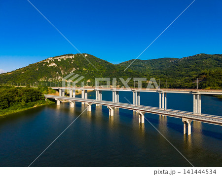 Bridge crosses a large body of water with green mountains in the background under a clear blue sky Bridge crosses a large body of water with green mountains in the background under a clear blue sky 141444534