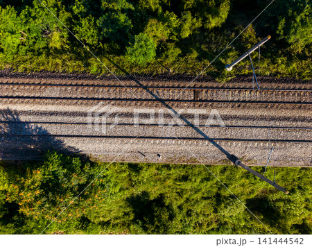 Train tracks run through wooded area near forest on a sunny day with clear skies and vibrant greenery around the sides of the path Train tracks run through wooded area near forest on a sunny day with clear skies and vibrant greenery around the sides of the path 141444542