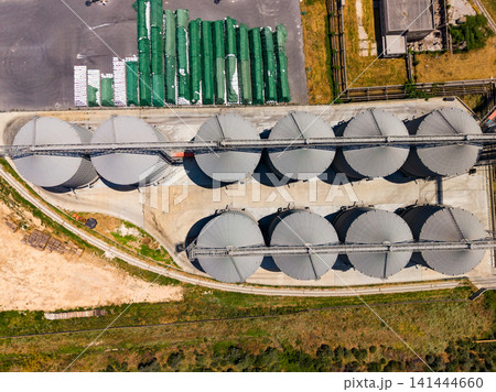 Large storage silos lined up on concrete, seen from above near a green agricultural area during daytime 141444660
