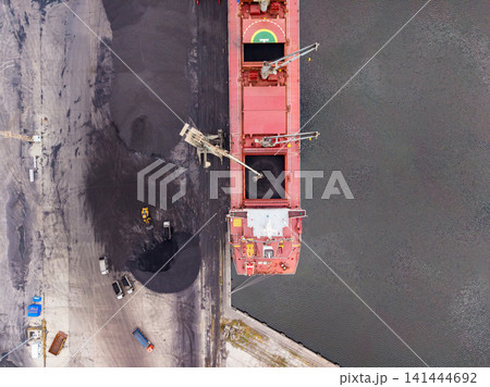 Large cargo ship unloads coal at a port while trucks drive nearby under a cloudy sky in the early afternoon Large cargo ship unloads coal at a port while trucks drive nearby under a cloudy sky in the early afternoon 141444692