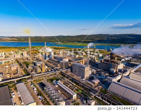 Industrial facility near water surrounded by hills with clear blue sky and clouds during daytime 141444723