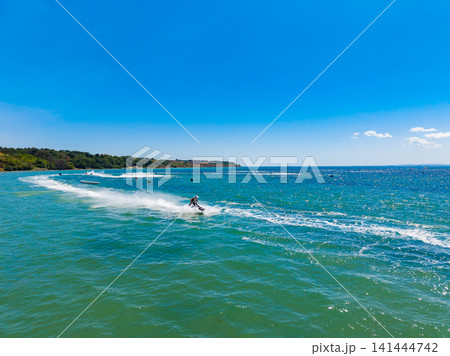 Surfers ride waves on a bright sunny day at a coastal location with clear blue skies and gentle surf Surfers ride waves on a bright sunny day at a coastal location with clear blue skies and gentle surf 141444742