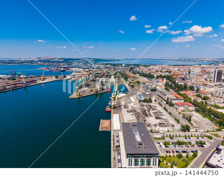 Overlooking a busy port area with cranes, ships, and a city skyline under a bright blue sky during daylight 141444750