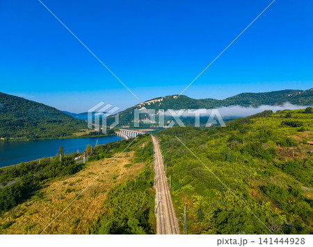 Train tracks run alongside a river and hills in a green landscape during a clear day Train tracks run alongside a river and hills in a green landscape during a clear day 141444928