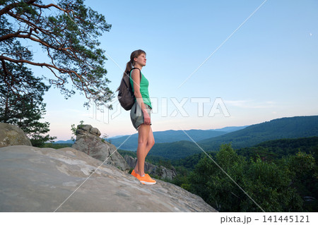 Hiker woman standing on mountain footpath enjoying evening nature during her travel on wilderness trail. Lonely female traveler traversing high hilltop route. Healthy lifestyle concept Hiker woman standing on mountain footpath enjoying evening nature during her travel on wilderness trail. Lonely female traveler traversing high hilltop route. Healthy lifestyle concept 141445121