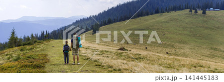 Father and son with backpacks hiking together in scenic summer green mountains. Dad and child standing enjoying landscape mountain view. Active lifestyle, family relations, weekend activity concept. Father and son with backpacks hiking together in scenic summer green mountains. Dad and child standing enjoying landscape mountain view. Active lifestyle, family relations, weekend activity concept. 141445183