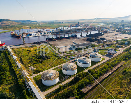 Cargo ships dock at a busy port with storage tanks and machinery in the foreground during the day 141445262