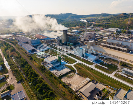 Factory complex with smoke in the sky during daylight hours near a rural area with green fields and gravel paths Factory complex with smoke in the sky during daylight hours near a rural area with green fields and gravel paths 141445267