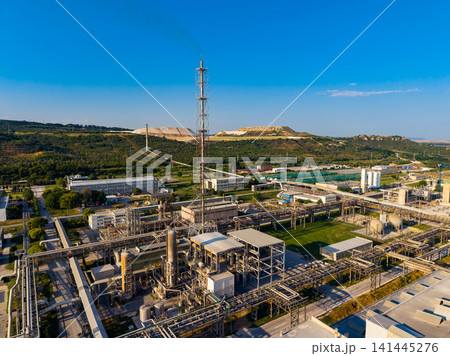 Large industrial plant with machinery, pipes, and towers located near hills under clear blue sky Large industrial plant with machinery, pipes, and towers located near hills under clear blue sky 141445276