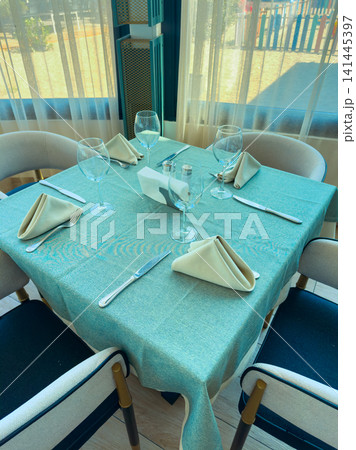 Dining table is set with cutlery and glassware in a cafe during the daytime near a beach area with sunlight streaming through the windows Dining table is set with cutlery and glassware in a cafe during the daytime near a beach area with sunlight streaming through the windows 141445397