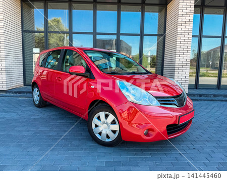 Bright red car parked in front of a modern building on a clear day with sunlight shining on the vehicle and surroundings Bright red car parked in front of a modern building on a clear day with sunlight shining on the vehicle and surroundings 141445460
