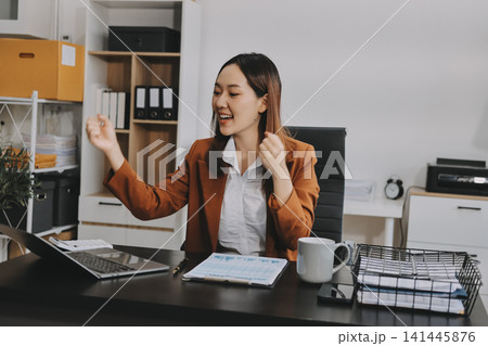 Young Asian professional business woman calculating finances and analyzing data charts on computer, smiling while working on accounting project in a modern office, planning budget 141445876