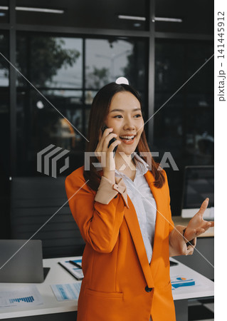 Smiling asian businesswoman talking on phone and using laptop while working at office desk 141445912