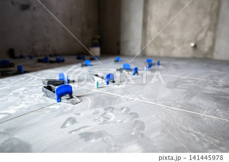 Grey floor tiles installing in a new house or apartment, showing clips and wedges of a tile leveling system ensuring an even surface during the renovation process Grey floor tiles installing in a new house or apartment, showing clips and wedges of a tile leveling system ensuring an even surface during the renovation process 141445978