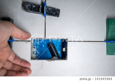 Hand placing ceramic tile around a blue electrical box, using a tile leveling system to ensure perfectly even surfaces during house renovation and construction 141445984