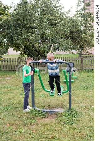 Two boys enjoying outdoor exercise equipment amidst green grass and trees, playing on a sunny day in a vibrant outdoor setting. 141446554