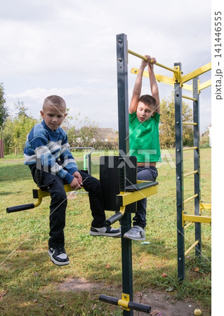 Two boys enjoying outdoor fitness equipment on a sunny day, one hanging on the bar, one sitting on the machine, focusing on their fitness. Two boys enjoying outdoor fitness equipment on a sunny day, one hanging on the bar, one sitting on the machine, focusing on their fitness. 141446555