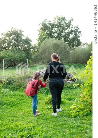 Mother and daughter holding hands outdoors, with a backpack, walking through a green, grassy landscape together on an adventure. Mother and daughter holding hands outdoors, with a backpack, walking through a green, grassy landscape together on an adventure. 141446663