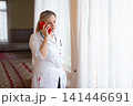 Female doctor on phone looking out window. Medical professional wearing white coat. Healthcare professional. Telemedicine concept. Patient care. 141446691
