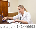 Professional woman in a white coat reviewing paperwork at a desk. She looks focused, wearing a stethoscope and holding a blue pen. 141446692