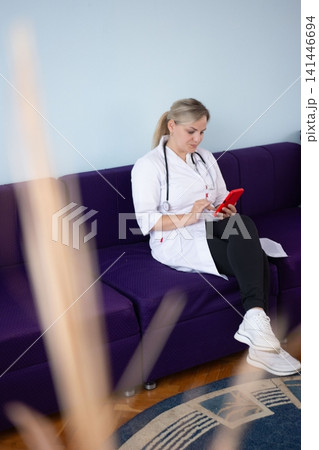 Doctor on a break, sitting on a purple couch, engrossed in her mobile phone. The scene evokes a moment of personal time in a professional setting. Doctor on a break, sitting on a purple couch, engrossed in her mobile phone. The scene evokes a moment of personal time in a professional setting. 141446694