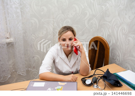 A healthcare provider is multitasking, conversing on a mobile phone while seemingly reviewing data on a computer at a light-colored desk. A healthcare provider is multitasking, conversing on a mobile phone while seemingly reviewing data on a computer at a light-colored desk. 141446698