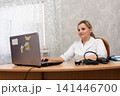Female doctor at her desk using a laptop, with medical equipment nearby, in a room with floral patterned wallpaper and sheer curtains. 141446700