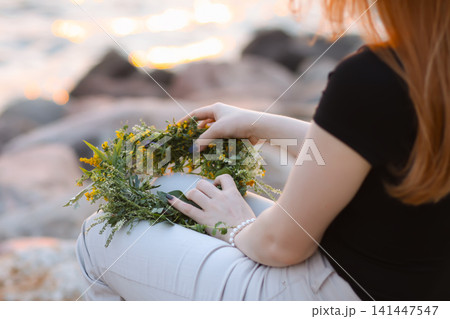 Woman resting outdoors in a natural summer landscape while holding a floral wreath made from meadow plants. 141447547