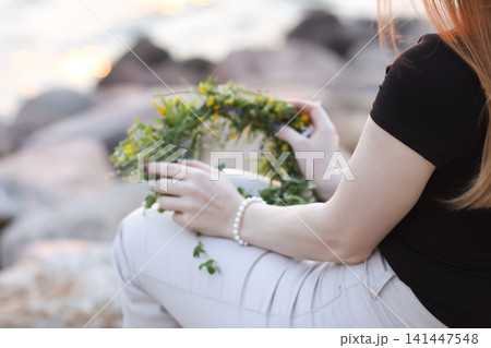 Young female holding a handcrafted flower and grass wreath while sitting among lush summer vegetation. Young female holding a handcrafted flower and grass wreath while sitting among lush summer vegetation. 141447548
