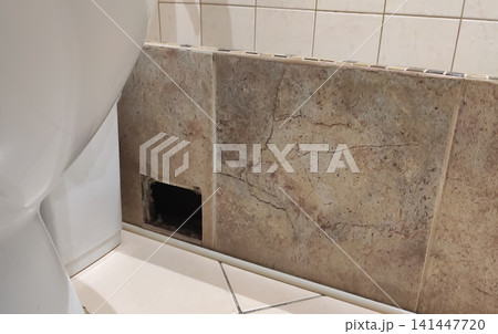 Damaged bathroom wall behind a toilet with missing tiles exposing a hidden hole in the surface. The defect may indicate water damage, plumbing issues, or the need for waterproofing inspection and Damaged bathroom wall behind a toilet with missing tiles exposing a hidden hole in the surface. The defect may indicate water damage, plumbing issues, or the need for waterproofing inspection and 141447720