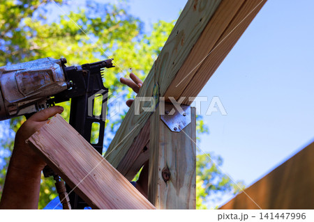 Hands hold air nail gun while securing wooden beams in construction project outdoors 141447996