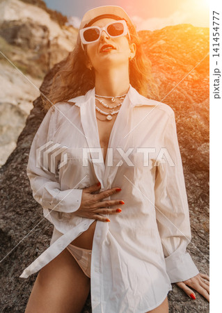 Woman summer beach posing in white shirt and sunglasses on rocky shore at sunset during vacation with copy space 141454777