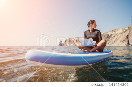 Woman sea laptop. Digital nomad, freelancer with laptop working on sup board at calm sea beach. Happy smiling girl relieves stress from work. Freelance, digital nomad, travel and holidays concept Woman sea laptop. Digital nomad, freelancer with laptop working on sup board at calm sea beach. Happy smiling girl relieves stress from work. Freelance, digital nomad, travel and holidays concept 141454802