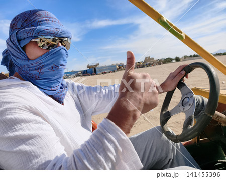 Desert thrill ride Arab man in traditional attire drives a dune buggy across the sun drenched sands, capturing the excitement of off road adventure under a clear blue sky. Desert thrill ride Arab man in traditional attire drives a dune buggy across the sun drenched sands, capturing the excitement of off road adventure under a clear blue sky. 141455396