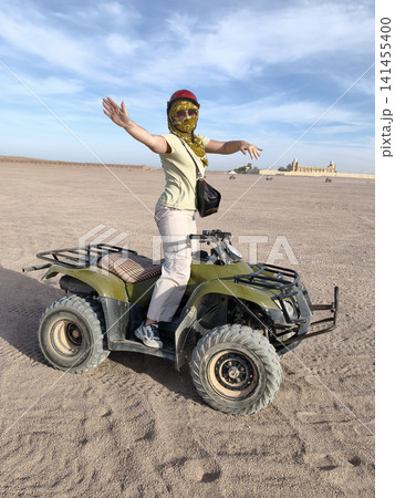 Adventurous woman casual summer outfit confidently rides green quad bike across golden desert dunes, arms raised in celebration, under a bright blue sky dynamic freedom, thrill outdoor exploration. Adventurous woman casual summer outfit confidently rides green quad bike across golden desert dunes, arms raised in celebration, under a bright blue sky dynamic freedom, thrill outdoor exploration. 141455400