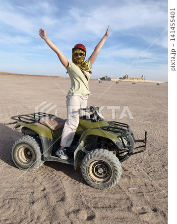 Adventurous woman casual summer outfit confidently rides green quad bike across golden desert dunes, arms raised in celebration, under a bright blue sky dynamic freedom, thrill outdoor exploration. Adventurous woman casual summer outfit confidently rides green quad bike across golden desert dunes, arms raised in celebration, under a bright blue sky dynamic freedom, thrill outdoor exploration. 141455401