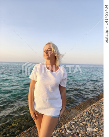 Joyful freedom by the sea carefree woman in crisp white top celebrates the moment with arms raised on a rocky shore, surrounded the calming rhythm of waves and the vast horizon under a clear blue sky 141455434