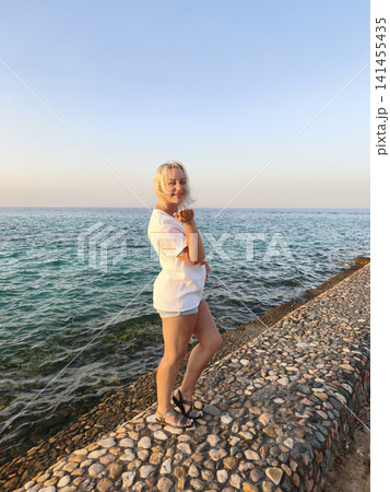 Joyful freedom by the sea carefree woman in crisp white top celebrates the moment with arms raised on a rocky shore, surrounded the calming rhythm of waves and the vast horizon under a clear blue sky 141455435