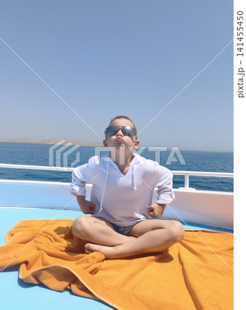 Young boy crisp white shirt and cool sunglasses lounges vibrant orange towel aboard sunlit deck endless blue sea and clear sky creating perfect backdrop for carefree summer days and joyful adventures 141455450