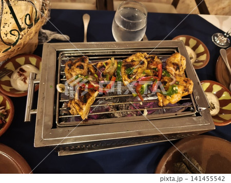 Grilled chicken pieces on a metal grill over glowing charcoal, showing appetizing char marks, juicy texture, and rich golden-brown color with smoke rising around. 141455542