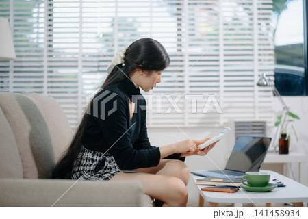 Young Asian woman managing home finances, calculating expenses, and paying bills with a credit card and laptop on a cozy sofa. 141458934
