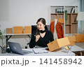 Asian online seller managing e-commerce business from home with laptop and shipping boxes. Live selling, social media marketing, order fulfillment and small business concept. 141458942