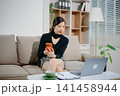 Young Asian businesswoman working remotely from home with laptop and coffee. Modern home office, freelance lifestyle, online business, productivity 141458944