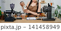 Focused Asian female barista taking stock and managing inventory with a clipboard at a modern cafe counter. 141458949